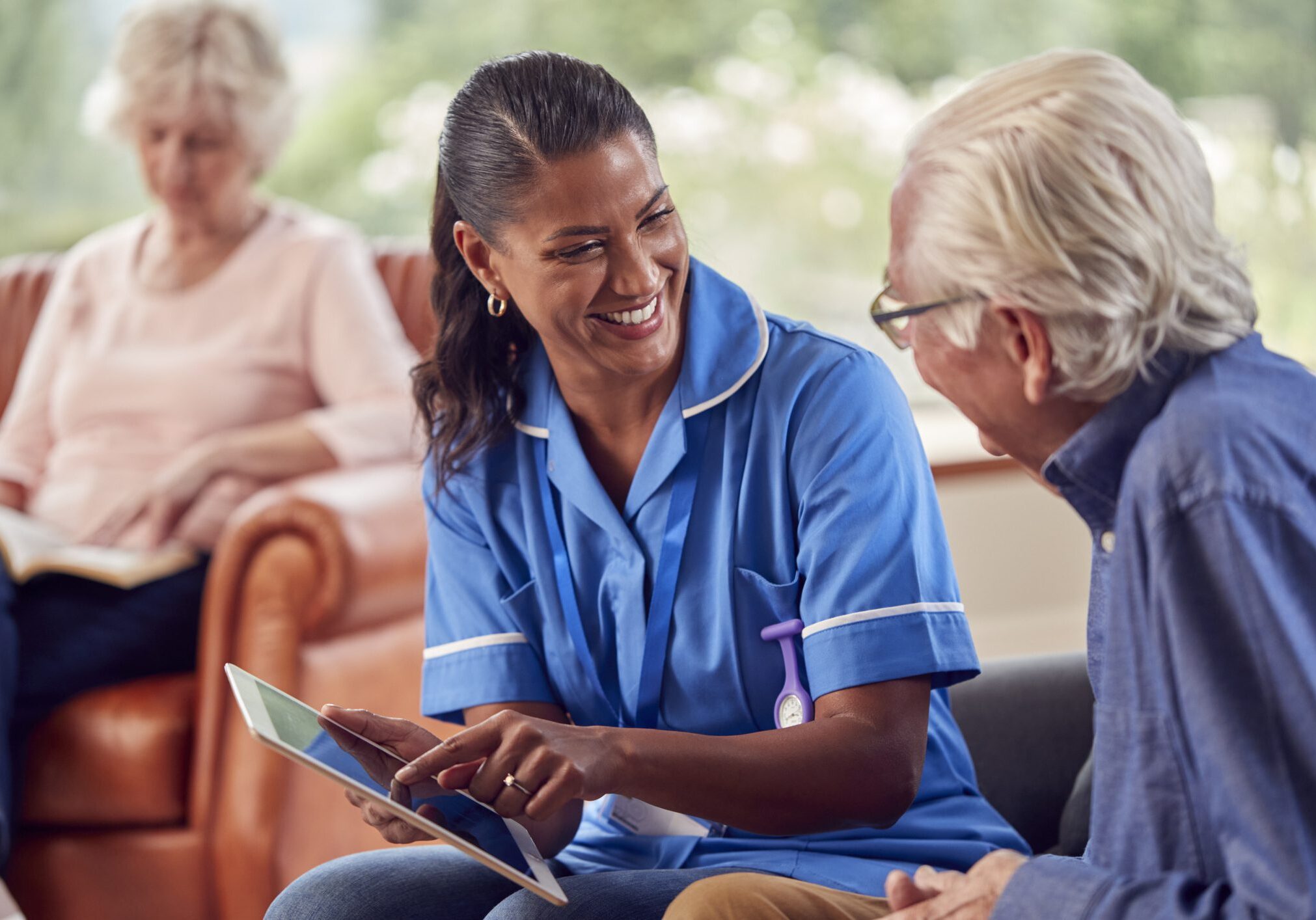 Senior Couple At Home With Man Talking To Female Nurse Or Care Worker Using Digital Tablet