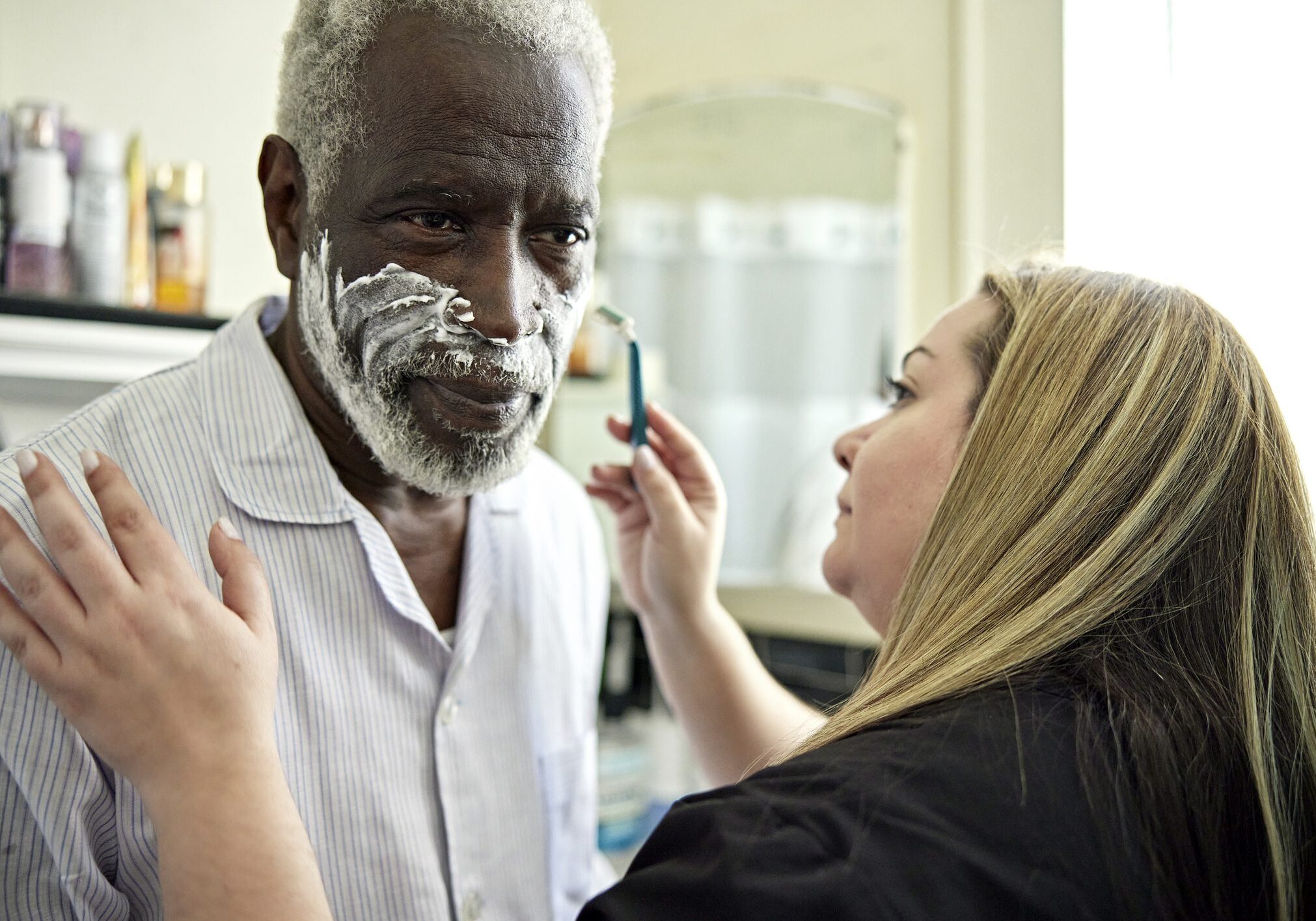 Head and shoulders view of Latin American woman in uniform assisting early 70s Black man with morning routine.